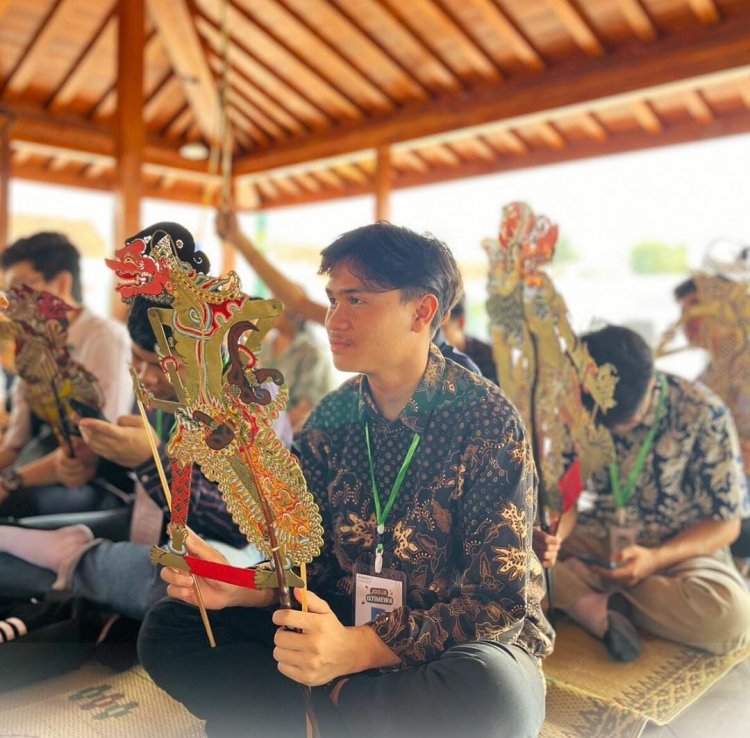 Seorang mahasiswa memegang wayang kulit dalam sesi pelatihan seni pedalangan, belajar tentang filosofi tokoh wayang dan teknik mendalang, dengan suasana pendopo yang penuh khidmat dan ornamen Jawa.